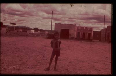 [Menino na praça de terra e casas]