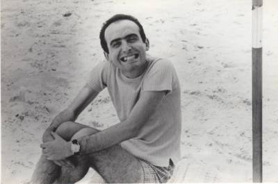 [Vladimir Herzog sentado na praia, abraçando os joelhos e sorrindo para a câmera]