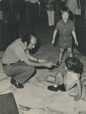 [Vladimir Herzog, agachado, aponta o dedo para Ivo Herzog criança sentado no chão]