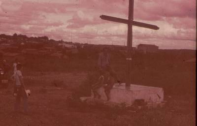 [Maria da Graça Soares Mascarenhas com chapéu na mão e Vladimir Herzog de pé sobre a base de uma cruz]